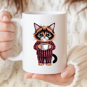 Peut inclure: Mug en céramique blanche avec un chat de dessin animé au regard renfrogné. Le chat porte un pyjama rayé rose et noir et tient une tasse de café. La tasse a une anse blanche et une finition brillante.