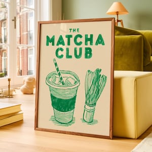 May include: A framed print with the text "THE MATCHA CLUB" in green. The image features a green drawing of an iced matcha drink with a straw and a matcha whisk. The frame is a dark wood colour.
