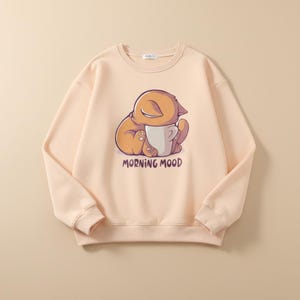 Peut inclure: Un sweat-shirt pêche clair avec un dessin de chat de dessin animé. Le chat, dans des tons orange et marron, serre une tasse blanche. Le texte "MORNING MOOD" est imprimé en dessous. Le sweat-shirt a un col rond.
