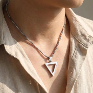 May include: A silver-coloured metal necklace featuring a box chain and a hollow triangular pendant. The pendant is a simple, open-frame triangle shape made of polished metal, suspended from a sturdy, rounded link chain that rests against the neck.