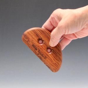 Puede incluir: Una herramienta de gua sha de madera con dos agujeros y el texto "Heim Tools" grabado en la superficie. La herramienta es marrón con un tono rojizo y tiene una forma curva.