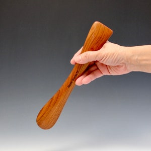 Könnte beinhalten: Ein Holzschuhspanner mit gebogenem Griff und abgerundetem Ende. Der Schuhspanner ist aus hellbraunem Holz gefertigt und trägt den eingravierten Text "Helm Tools" auf der Seite.