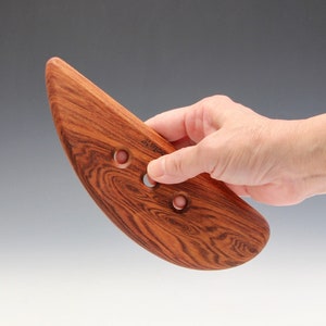 Puede incluir: Una herramienta de costilla de cerámica de madera con forma de lágrima curvada. La herramienta está hecha de madera de color marrón rojizo con un patrón de vetas prominente y tres agujeros circulares. La herramienta se sostiene en una mano.