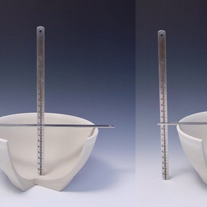May include: A white ceramic bowl with a unique, asymmetrical design. The bowl is measured with a ruler, showing its dimensions.