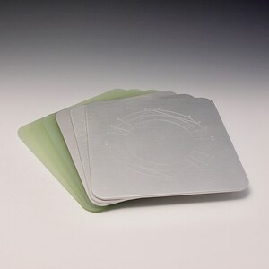 May include: A stack of five light green and silver square-shaped coasters with a circular design on the top. The design includes a circle with lines radiating outward.