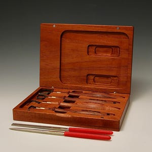 May include: A set of six wood carving tools with wooden handles and metal blades, in a wooden box with a hinged lid. The tools are arranged in the box, and there are two red-handled files next to the box.