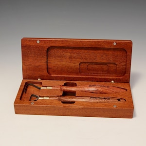 May include: Two wooden handled tools with brass tips in a wooden box. The box is made of mahogany wood and has a hinged lid. The tools are labeled "Helm Tools".