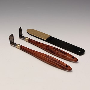 Può includere: Tre strumenti per artisti: due spatole con manici in legno e lame in metallo, e un bastoncino di levigatura nero con una superficie abrasiva color oro. Le spatole hanno ghiere in ottone e il testo "Hsin Book" sui manici.