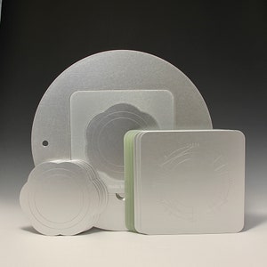 May include: A set of silver metal cake decorating stencils. The set includes a large round stencil, a smaller round stencil with a flower design, and a square stencil with a circular design. The stencils are stacked on top of each other.