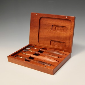 May include: A set of five wood carving tools with wooden handles in a wooden box. The tools are arranged in the box with the handles facing up. The box is open and the tools are visible.