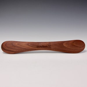 Könnte beinhalten: Ein Holzlöffel mit einem abgerundeten Griff und einer leicht gekrümmten Schale. Der Löffel ist aus Walnussholz gefertigt und trägt die Worte "New Woods" auf dem Griff.