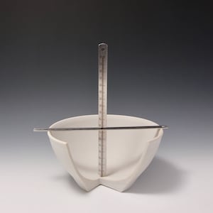 May include: A white ceramic bowl with a unique, angular design. The bowl is split in half and has a ruler measuring 15 cm in height placed in the center.