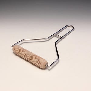 May include: A decorative paint roller with a light brown patterned roller and a silver metal handle. The roller features a repeating geometric design. The handle is a simple, ergonomic design.