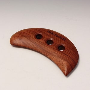 May include: A crescent-shaped wooden tool with three holes, made for massage. The tool is brown and has the text "Heal Tools" engraved on it.