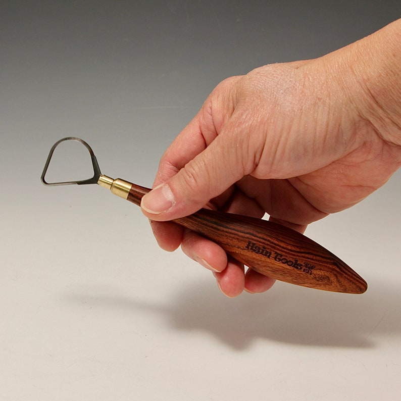 May include: A close-up of a wooden pottery tool with a metal loop at the end. The tool is held in a hand and has the words "Hein Tools" engraved on the side.