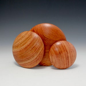 Könnte beinhalten: Drei Holzschalen in verschiedenen Größen, alle in einem rötlich-braunen Farbton mit einer Holzmaserung. Die Schalen sind versetzt angeordnet, wobei die größte Schale hinten und die kleinste Schale vorne ist.