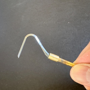 May include: A metal dental scaler tool featuring a curved, pointed silver-colored tip and a cylindrical gold-toned handle with a threaded end. The instrument has a smooth, reflective finish on the hook portion and a textured grip on the handle.