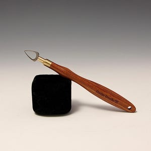May include: A wood-handled clay sculpting tool with a curved blade and brass ferrule. The handle is a reddish-brown color, and the tool rests on a black square. The text "Hein Tools" is printed on the handle.
