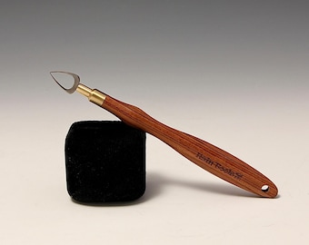 May include: A wood-handled clay sculpting tool with a curved blade and brass ferrule. The handle is a reddish-brown color, and the tool rests on a black square. The text "Hein Tools" is printed on the handle.