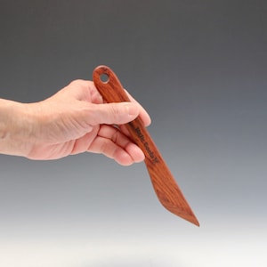 May include: A wooden letter opener with a curved blade and a handle with a hole. The letter opener is made of dark wood and has the words "Wood Tools" engraved on the blade.