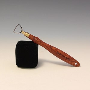 May include: A wood carving tool with a small, curved blade and a wooden handle. The handle is made of reddish-brown wood and has the words "West Tools" engraved on it.