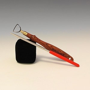 May include: A set of two wood carving tools with red handles. The tools are on a black velvet surface. The larger tool has a wooden handle with the text "Hein Tools" engraved on it. The smaller tool has a red handle and a pointed tip.