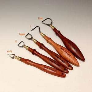 May include: A collection of five pottery tools with wooden handles and metal blades. The tools have different blade shapes, including arch, D, square, and pear. The handles are a dark brown color, and the blades are silver.