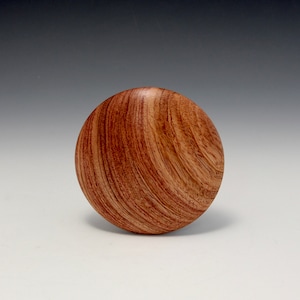 May include: A circular wooden object in shades of reddish-brown. The surface has a swirling grain pattern, giving it a textured look. It could be a decorative item or part of a larger piece, possibly a lid or knob.