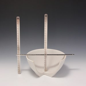 May include: A white ceramic bowl with a unique, asymmetrical shape. The bowl is measured with three metal rulers.