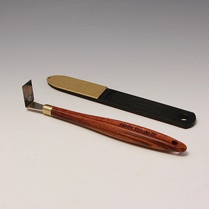 Può includere: Un coltello di precisione con manico in legno, raccordo in ottone e lama angolata, insieme a una pietra per affilare nera in un astuccio. Il manico del coltello è inciso con la scritta "Hain Tools".