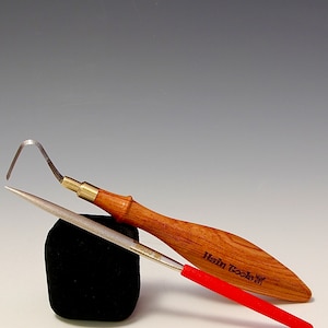 May include: Two woodworking tools are displayed against a gradient background. One tool has a curved blade and a wooden handle with brass accents. The other is a metal file with a red handle. A black foam block supports the tools.