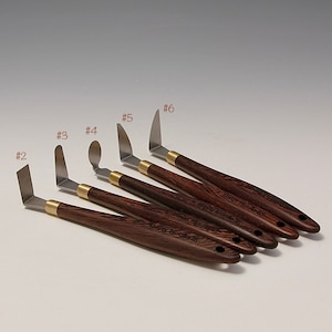 Può includere: Set di sei coltelli da tavolo in acciaio inossidabile con manici in legno. I coltelli sono numerati da 2 a 6 e hanno una varietà di forme, tra cui una quadrata, una punta arrotondata e una punta appuntita.