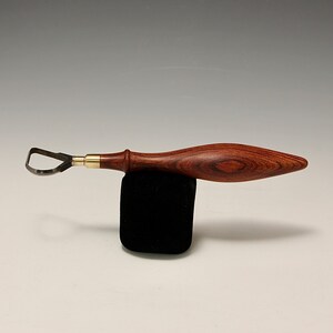 May include: A wood carving tool with a curved blade and a wooden handle. The handle is made of dark wood with a reddish brown colour.