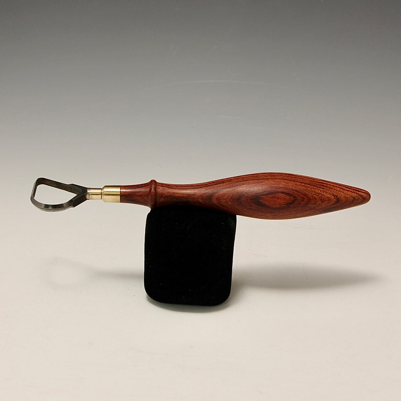 May include: A wood carving tool with a curved blade and a wooden handle. The handle is made of dark wood with a reddish brown colour.
