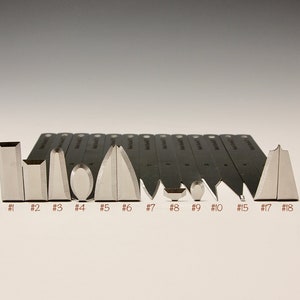 Puede incluir: Un conjunto de 13 cuchillas de metal con diferentes formas y tamaños, etiquetadas con números del 1 al 18. Las cuchillas están dispuestas en una fila sobre una superficie blanca.