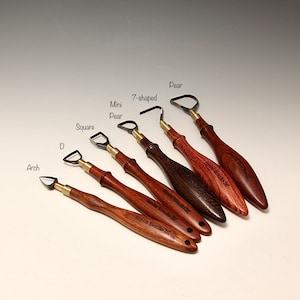 May include: A collection of six wood-handled clay sculpting tools. Each tool has a different shaped metal loop or blade at the end, including Arch, D, Square, Mini Pear, 7-shaped, and Pear. The handles are a rich, reddish-brown color.