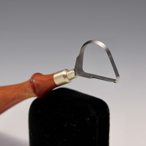May include: A carving tool with a wooden handle and a metal loop-shaped blade. The tool is resting on a black surface.