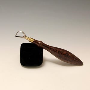 May include: A wood-handled pottery rib tool with a curved metal blade and brass ferrule. The handle features a dark brown, patterned wood grain. The tool rests on a small, black, velvet-covered block.
