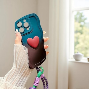 Könnte beinhalten: Eine teal- und braunfarbene Handyhülle mit einem roten Herzdesign. Die Hülle hat einen Farbverlauf und einen Riemen mit grünen, lila und weißen Akzenten. Die Hülle soll das Telefon schützen und eine dekorative Note verleihen.