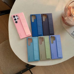 May include: A collection of colorful phone cases with attached grip stands. The cases are in various shades, including pink, blue, green, and purple. Each case has the text "GripCase" printed on it.