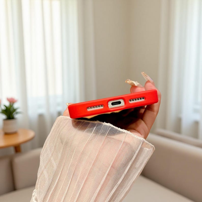 Puede incluir: Una funda de tel&eacute;fono roja brillante con recortes para la c&aacute;mara, el puerto de carga y los altavoces. La funda se sostiene en una mano, con una manga de encaje blanco transparente en primer plano. El fondo est&aacute; borroso, con un vistazo a una planta y muebles.