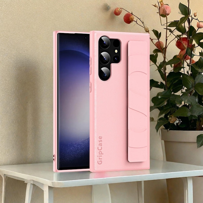 May include: A light pink phone case with a built-in grip and camera cutouts. The case is shown with a phone inside, and the text "GripCase" is printed vertically on the side. The phone screen displays a blue and white gradient.