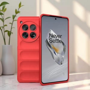 Op de afbeelding: Rode telefoonhoes met een beschermende cameralensafdekking en de tekst "Never Settle" op het scherm. De hoes heeft een getextureerde achterkant en een verhoogde rand voor schermbescherming. De telefoonhoes wordt getoond naast een plant in een vaas.
