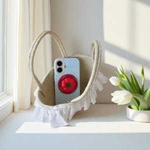 May include: A light blue phone case featuring a red tomato design sits in a beige woven basket with white lace trim. The basket has a handle. A vase of white tulips is to the right. The basket is on a white surface.