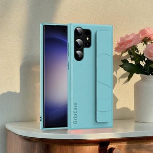 May include: A light blue phone case with the text "GripCase" is shown next to a smartphone. The case has a textured surface and a built-in grip. The phone's screen is visible. A vase of pink roses is in the background.