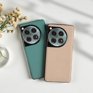 May include: Two phone cases, one teal and one beige, with a textured leather-like finish. Each case has a black camera module with four lenses. The cases are displayed on a white surface with a small vase of flowers.