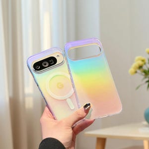 Puede incluir: Dos fundas transparentes para teléfono con un diseño de degradado arcoíris. Una funda está adherida a un teléfono, mostrando la cámara y un área de carga magnética circular. La otra funda está vacía. Las fundas tienen un borde transparente con un tono púrpura.