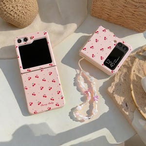 May include: Two pink phone cases adorned with a cherry pattern. One is open, showing a black screen, and the other is closed, displaying a clock. A beaded phone charm with pink and white beads and a pink bow is attached to the closed case. The text "Cherry Baby" is visible.
