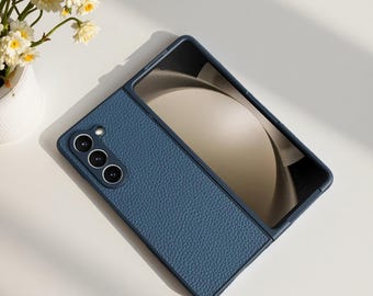 Puede incluir: Una funda de teléfono azul marino con un acabado texturizado similar al cuero. La funda está abierta, revelando la pantalla del teléfono. El teléfono tiene tres lentes de cámara. La funda está sobre una superficie blanca con un pequeño jarrón de flores.