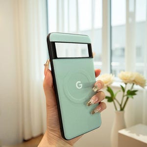 May include: A mint green phone case with a black frame, featuring a circular design with the letter "G" in the center. The case has a rectangular cutout at the top. The case is held in a hand.